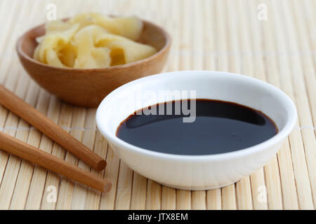 La sauce soja dans un bol blanc sur le tapis de bambou avec des baguettes et gingembre mariné dans bol en bois. Banque D'Images