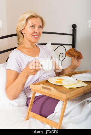 Smiling blonde mature woman enjoying petit-déjeuner au lit avec croissant et café Banque D'Images