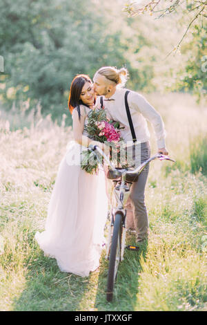 La jolie photo de la couple transportant la bicyclette et la marche dans la forêt ensoleillée. Le marié est habillé moderne s'embrasser la mariée dans les longs cheveux blancs dans la joue. Banque D'Images