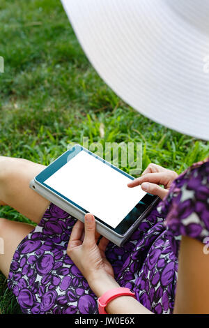 Femme anonyme dans white hat et robe de détente sur l'herbe verte avec tablette. Vue arrière. Petit coup à orientation verticale modèle de vue. Banque D'Images