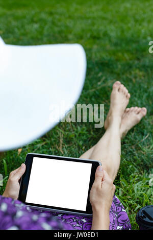 Femme anonyme dans white hat et robe de détente sur l'herbe verte avec tablette. Vue arrière. Petit coup avec écran en position horizontale, le modèle. Banque D'Images