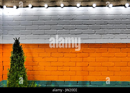 Petit arbuste contre mur en brique avec blanc, orange et vert bandes horizontales et chaîne de lumières au-dessus Banque D'Images