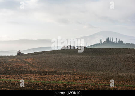 Les tracteurs modernes sur la Toscane gris terre labourée et distant des cyprès autour d'une villa, disparaissant dans la brume Banque D'Images
