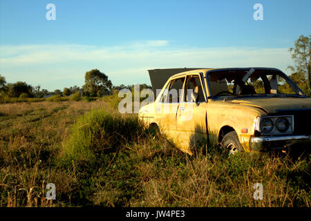 Une vieille voiture jaune abandonné et négligé de gauche à la rouille dans un champ sur une ferme avec les mauvaises herbes qui poussent autour d'elle. Banque D'Images