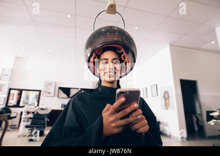 Femme au spa avec des rouleaux à ses cheveux et une machine à vapeur autour de la tête. Smiling woman looking at le téléphone mobile au salon de beauté tout en Banque D'Images