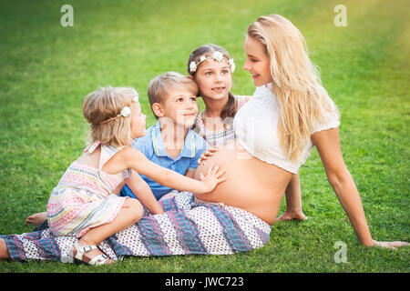 Jeune mère enceinte assis sur l'herbe verte fraîche champ avec ses trois enfants, les enfants sont impatients de la naissance d'un frère ou soeur, h Banque D'Images