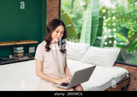 Beau young smiling woman working on laptop while sitting on bed at home près de grande fenêtre. Banque D'Images