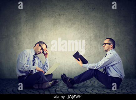 Par rapport à l'ordinateur d'adresses. Deux hommes assis sur un plancher l'un de lire un livre un autre working on laptop Banque D'Images