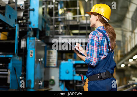 Portrait of female factory worker writing on clipboard lors de la vérification de l'utilisation de machines dans une usine moderne Banque D'Images