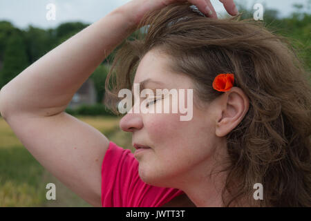 Une femme dans un haut rose tirant ses cheveux en arrière pour révéler un champ sauvage coquelicot derrière son oreille sur une journée d'été. UK Banque D'Images