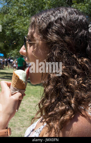 Belle jeune femme aux cheveux bruns frisés eating ice cream au Festival Austin Ice Cream fond pittoresques en plein air avec des foules et des parasols. Banque D'Images