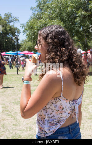 Belle jeune femme aux cheveux bruns frisés eating ice cream au Festival Austin Ice Cream fond pittoresques en plein air avec des foules et des parasols. Banque D'Images