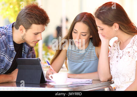 Trois étudiants concentré à travailler ensemble et prendre des notes en ligne assis sur table d'une terrasse de restaurant Banque D'Images