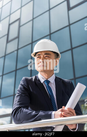 Portrait of pensive architecte professionnel in hard hat holding blueprint Banque D'Images