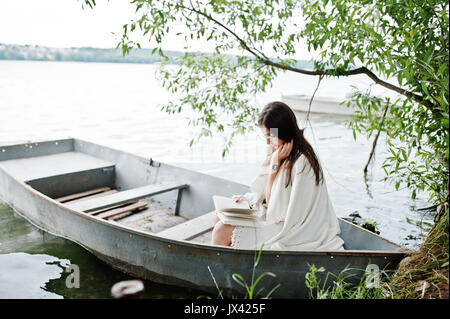 Portrait d'une jolie femme portant des pois noir robe, châle blanc et verres de lire un livre dans un bateau sur un lac. Banque D'Images