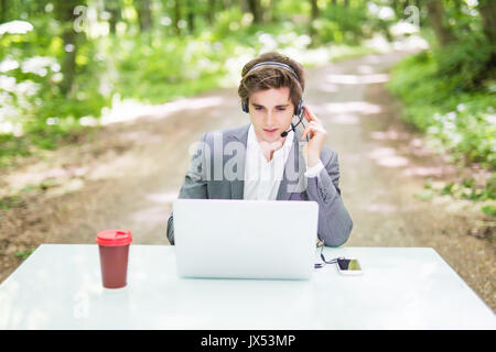 Handsome Businessman with headset sitting au bureau bureau avec ordinateur portable et tasse de café au centre d'appels à Green Forest Park . Banque D'Images