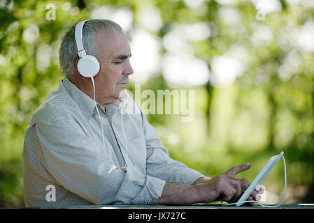 Un homme âgé d'écouter de la musique sur un ordinateur tablette assis à l'ombre reposante des arbres dans le parc en sélectionnant une mélodie sur l'écran tactile Banque D'Images