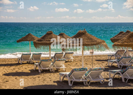 D'une manière ordonnée de bordée de transats recouverts de tissu floral lumineux, sous des parasols de paille pour attendre les touristes sur la Costa del Sol, Espagne Banque D'Images