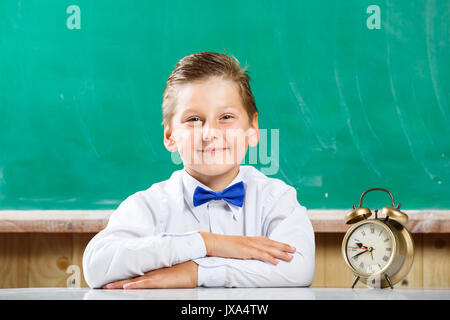 Petit garçon en chemise assis avec réveil près de tableau noir. Peu de livres avec des élèves en attente de leçons à l'école Banque D'Images