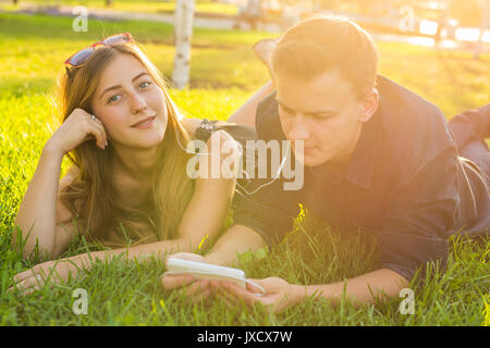 Portrait ensoleillée de sweet young couple lying détente sur l'herbe et écoute de la musique des écouteurs sur smartphone, journée d'été Banque D'Images