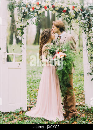 Le close-up photo verticale du marié en costume d'époque s'embrasser son épouse avec le grand bouquet dans le front sous l'arche dans la forme du livre blanc deux portes décorées avec les fleurs colorées. L'emplacement des forêts. Banque D'Images