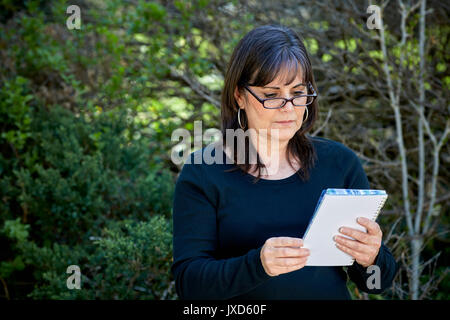Belle femme d'âge moyen portant des lunettes de lecture en regardant le cahier dans ses mains en plein air Banque D'Images