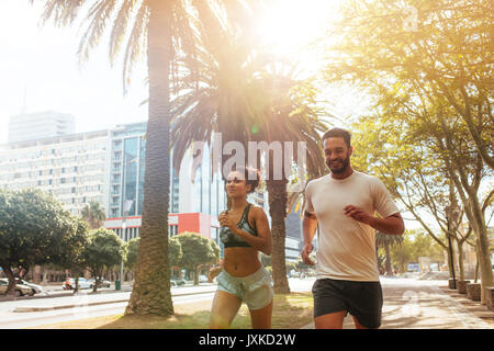 Jeune homme et femme en tenue de jogging courir ensemble à l'extérieur. Couple d'exécution sur rue avec arbres et bâtiments en arrière-plan. Banque D'Images