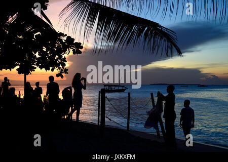 Tanzanie Zanzibar, Stone town, Livingstone beach restaurant and bar, soirée en mer, touriste étranger en bar, femme de prendre une photo de coucher de soleil et colporteur vente de vêtements Banque D'Images