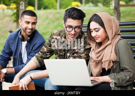 Trois amis, assis sur un banc de parc, looking at laptop Banque D'Images