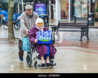 Vieux couple shopping sous la pluie, l'un poussant l'autre dans un fauteuil roulant, transportant un sac Tesco 'pour la vie'. 'Le petit sac vert'. Vieux mobilité de la pluie. Banque D'Images