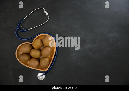 High angle photo de beaucoup de délicieux kiwis en plaque en bois en forme de cœur montrant sur tableau noir avec stéthoscope médical. Banque D'Images