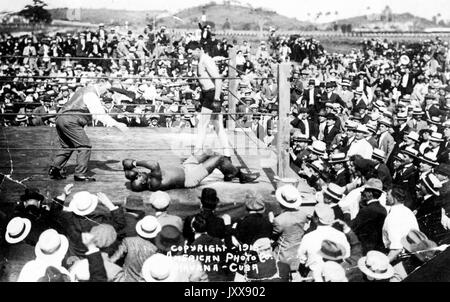 Un grand groupe de spectateurs se tient autour d'un ring de boxe en plein air, dans lequel sont Jack Johnson (à droite), un arbitre (à gauche), et Jess Willard (au sol), qui vient d'être frappé par Johnson au 26e tour pour gagner le championnat mondial de boxe poids lourds, la Havane, Cuba, 1915. Banque D'Images