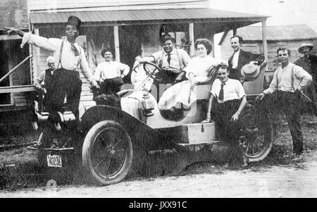 Un groupe de personnes, dont huit hommes et deux femmes, souriant pour la plupart, se tiennent debout sur ou autour d'une voiture, avec un homme afro-américain portant un tophat debout sur l'aile et pointant avec son bras tendu, 1910. Banque D'Images