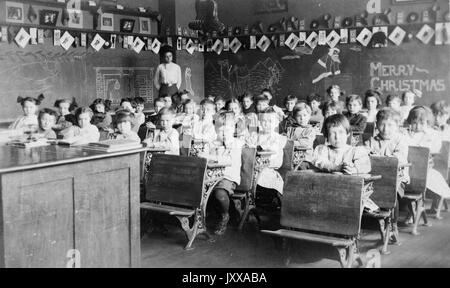 Photo de paysage pleine longueur d'écoliers assis à des bureaux, professeur afro-américain à l'arrière de la salle, « Joyeux Noël » écrit sur le tableau noir, 1920. Banque D'Images