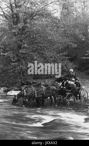 Homme afro-américain en tant qu'entraîneur de calèche conduit par deux chevaux portant deux femmes blanches, suivi d'un homme blanc à cheval, d'un homme afro-américain portant un costume sombre, d'une femme blanche portant une robe et un chapeau clairs, d'une autre femme blanche portant une robe et un chapeau foncés, homme blanc dans le dos portant une chemise légère, sur la voie de la terre devant les arbres, expressions neutres, 1915. Banque D'Images