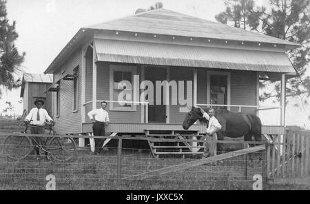 Portraits sur pied de deux hommes blancs matures et d'un homme afro-américain, un homme blanc portant un bouton léger, une cravate et un pantalon sombre, debout à côté d'un cheval, un autre homme blanc se penchant contre la maison, portant un bouton blanc et un pantalon sombre, un homme afro-américain portant un bouton léger, pantalon et chapeau sombres, debout derrière un vélo, tous debout devant la maison, expressions neutres, 1920. Banque D'Images