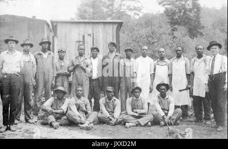 Un groupe de jeunes Afro-américains matures aux expressions neutres se tiennent debout et assis à l'extérieur de bâtiments banteux, avec deux hommes caucasiens (à l'extrême gauche et à l'extrême droite) vêtus de vêtements plus fins, qui semblent être leurs supérieurs, 1915. Banque D'Images