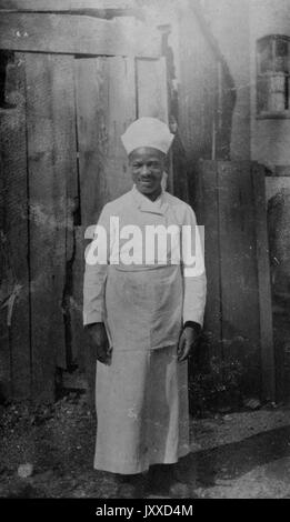 Portrait complet d'un homme afro-américain, habillé d'un tablier et d'un chapeau de chef ou de marin, dans une cuisine, expression faciale souriante, 1920. Banque D'Images