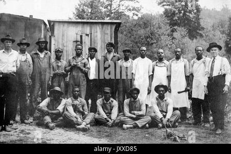 Un groupe de travailleurs afro-américains aux expressions neutres, certains vêtus de combinaisons tandis que d'autres portent des tabliers, rassemblés à l'extérieur dans une zone boisée dégagée, avec deux hommes blancs vêtus de vêtements plus fins qui se tiennent de part et d'autre du groupe et semblent être les supérieurs des travailleurs, 1915. Banque D'Images