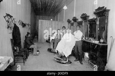 Un groupe d'Afro-Américains, dont deux patrons masculins, un jeune homme qui attend une chaise, un jeune homme qui barber et une femme, sont rassemblés dans un salon de coiffure, contenant des chaises, des porte-chapeaux et des porte-manteaux, et trois postes avec des chaises, des miroirs et du matériel pour barbiers, 1912. Banque D'Images