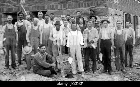 Un groupe de jeunes et matures travailleurs américains aux expressions neutres se tient devant un bâtiment où sont livrés des outils, dont des bêches, un marteau et une canette à pétrole, 1925. Banque D'Images