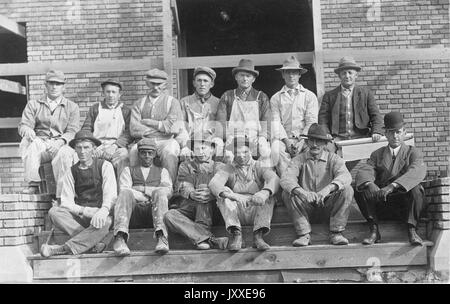 Groupe de jeunes et matures travailleurs américains aux expressions neutres assis sur des marches à l'extérieur d'un bâtiment, avec un homme (en bas à droite) habillé de vêtements plus fins, y compris un chapeau sophistiqué et des gants en cuir, qui semble être leur supérieur, 1920. Banque D'Images