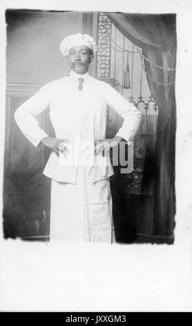 Portrait de trois quarts de longueur d'un homme afro-américain mature, portant un ensemble léger, une chemise claire, une cravate sombre et un chapeau de chef, tenant un cigare dans la bouche, debout devant la toile de fond, expression neutre, 1920. Banque D'Images