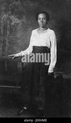 Portrait d'une femme afro-américaine debout devant une murale et appuyée contre une chaise en bois, son bras droit repose également sur une autre chaise en bois, son bras gauche est à côté, elle porte une longue jupe de couleur foncée et un chemisier de couleur claire, 1915. Banque D'Images