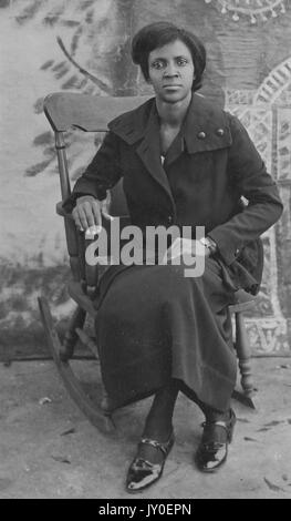 Portrait complet d'une femme afro-américaine mûre avec expression neutre devant un fond peint, assise sur une chaise à bascule en bois, portant un manteau, une jupe et des chaussures sombres, 1925. Banque D'Images