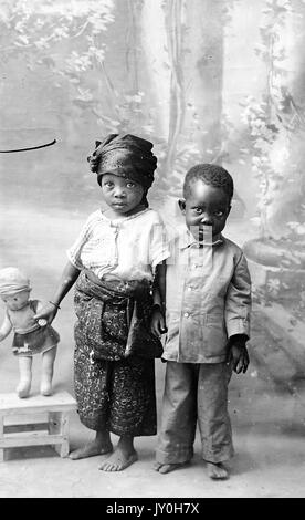 Portrait debout de deux enfants afro-américains, fille portant une chemise claire avec un pantalon à motifs foncés et un serre-tête, fille portant une poupée bébé, garçon portant une chemise et un pantalon foncés, debout devant la toile de fond, expressions neutres, 1920. Banque D'Images