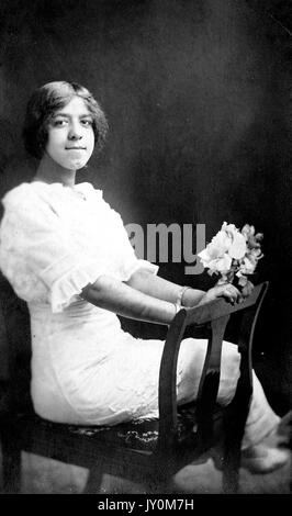 Portrait intégral d'une jeune afro-américaine assise sur une chaise en bois, portant une robe blanche, portant un petit bouquet de fleurs, les deux mains pressées sur l'arrière de la chaise, avec une expression faciale sérieuse, 1920. Banque D'Images