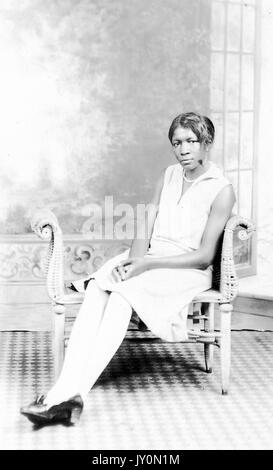 Portrait complet d'une femme afro-américaine assise, portant une robe blanche et des bas blancs, les mains repliées sur ses genoux, portant un collier de perles, assise dans une chaise en bois devant la toile de fond, 1920. Banque D'Images