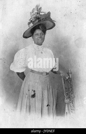 Portrait à trois quarts d'une femme afro-américaine debout à côté d'une chaise, avec une main posée sur la chaise, portant une robe à carreaux et un chemisier blanc, avec un chapeau à plumes orné sur sa tête, avec une expression faciale souriante, 1920. Banque D'Images