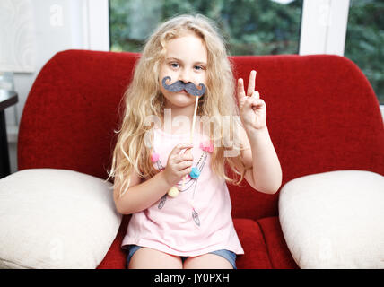 Jolie petite fille avec des moustaches de papier alors qu'il était assis sur le fauteuil rouge à la maison. Banque D'Images